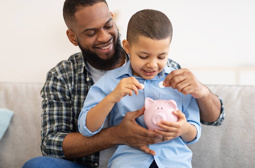 Em uma sala um pai negro, de barba e com uma blusa xadrez, abraçado com seu filho colocam uma moeda em um cofre no formato de um porquinho rosa.