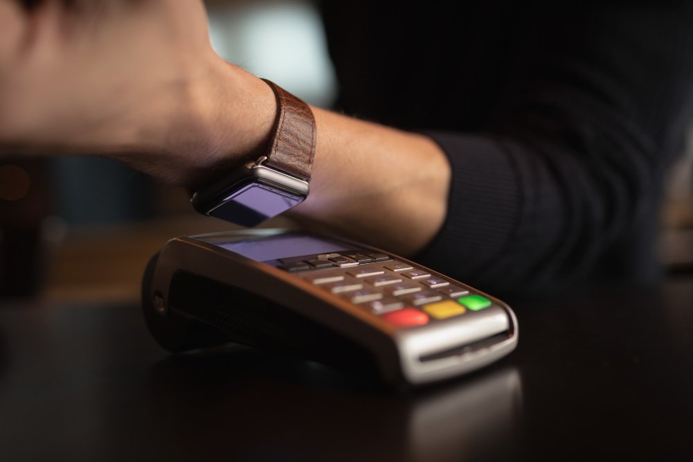 Close em um braço de um homem branco usando uma blusa preta, aproximando o smartwatch de uma máquina de cartões